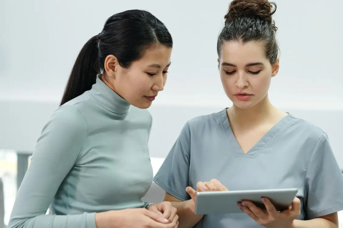 Two women talking to each other, one is wearing a scrub suit while holding a tablet, and the other is wearing a casual clothing at Synapse Revenue Cycle Management.